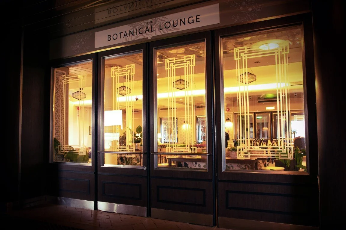 Entrance door to 'The Botanical Lounge' restaurant with glass panels, gold decorative patterns, and warm interior lighting visible through the windows.