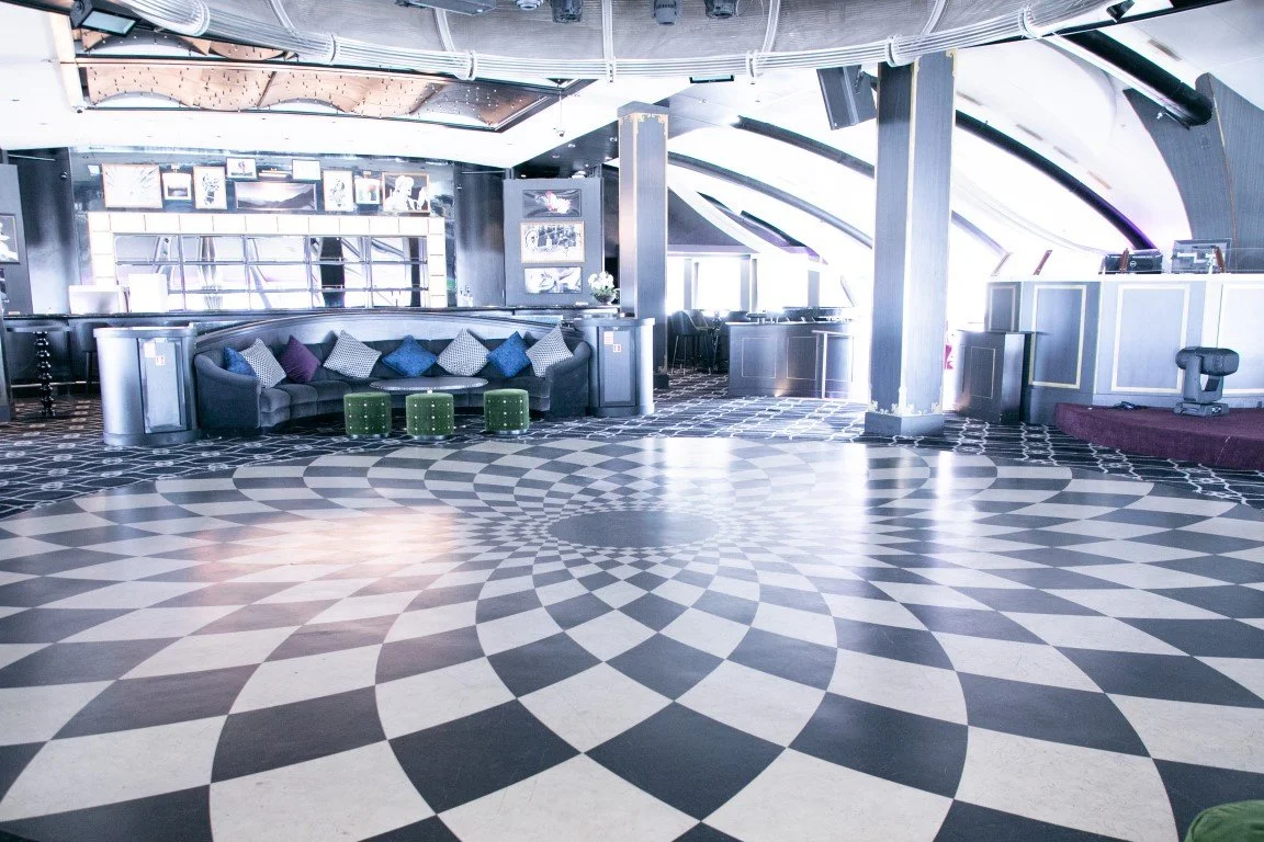 Empty modern lounge with patterned black and white floor, a curved black sofa with colorful pillows, a bar area, and a DJ booth at the back, in a venue with high ceiling and large windows.