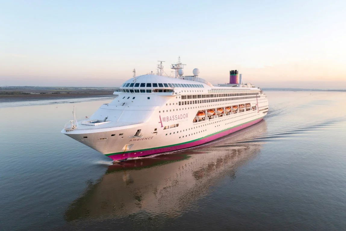 A large white cruise ship named 'Ambiance' sailing on calm water at sunset, with an open deck on the side showing orange lifeboats.