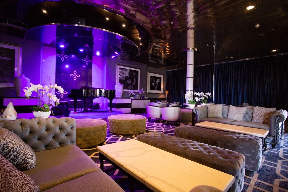 Luxurious lounge with modern furniture, purple lighting over a small stage with a black grand piano, and floral arrangements on tables.