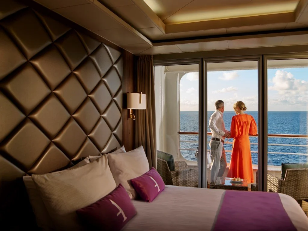 A hotel room with a large bed, beige pillows, and purple pillows, with a balcony door open to a view of the ocean and two people holding hands on the balcony.