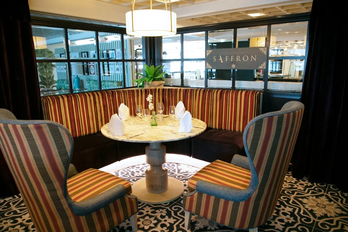 A round table with four glasses and napkins set for dining, surrounded by two upholstered chairs and a curved bench with striped fabric, in a cozy restaurant corner with a sign that says 'Saffron' behind a glass partition.
