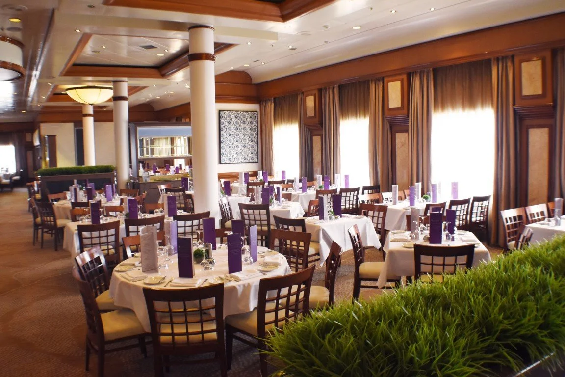 Elegant restaurant with round tables covered in white tablecloths, purple menus, and place settings. Large windows with heavy curtains, wooden accents, and lush green plants create a sophisticated atmosphere.