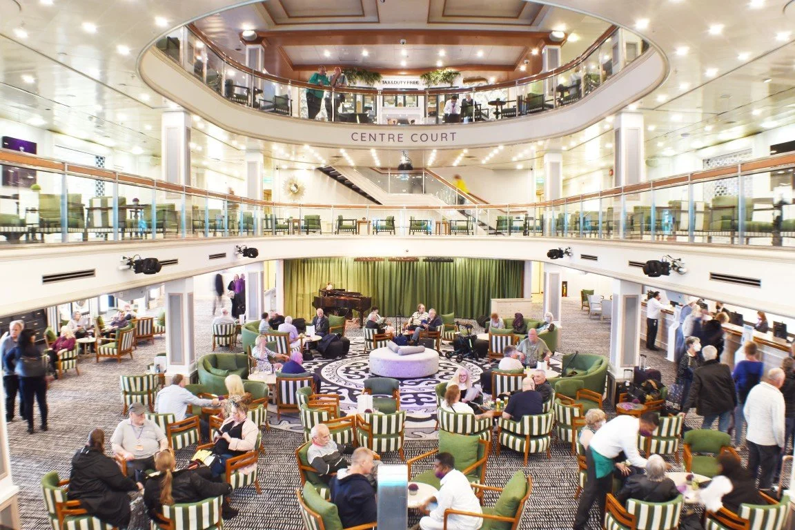 Interior of a multi-level shopping mall or hotel lobby with seating area, a stage with a piano, green curtains, and multiple people strolling, sitting, and talking.