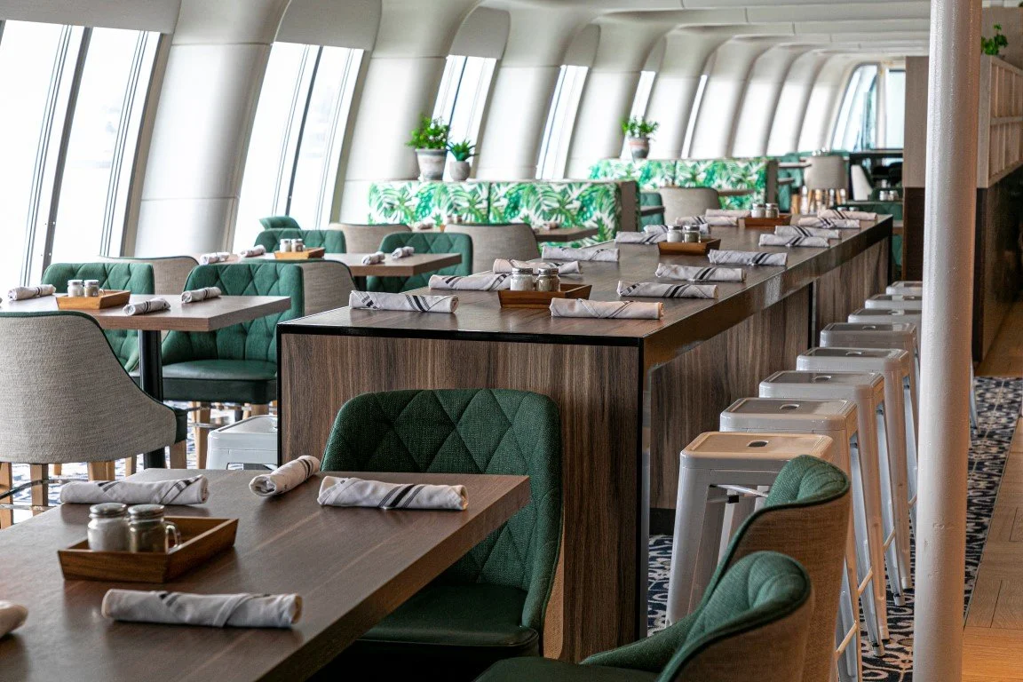 A modern restaurant interior with wooden tables and green upholstered chairs, set with napkins and cutlery, near large windows in a high-rise building.