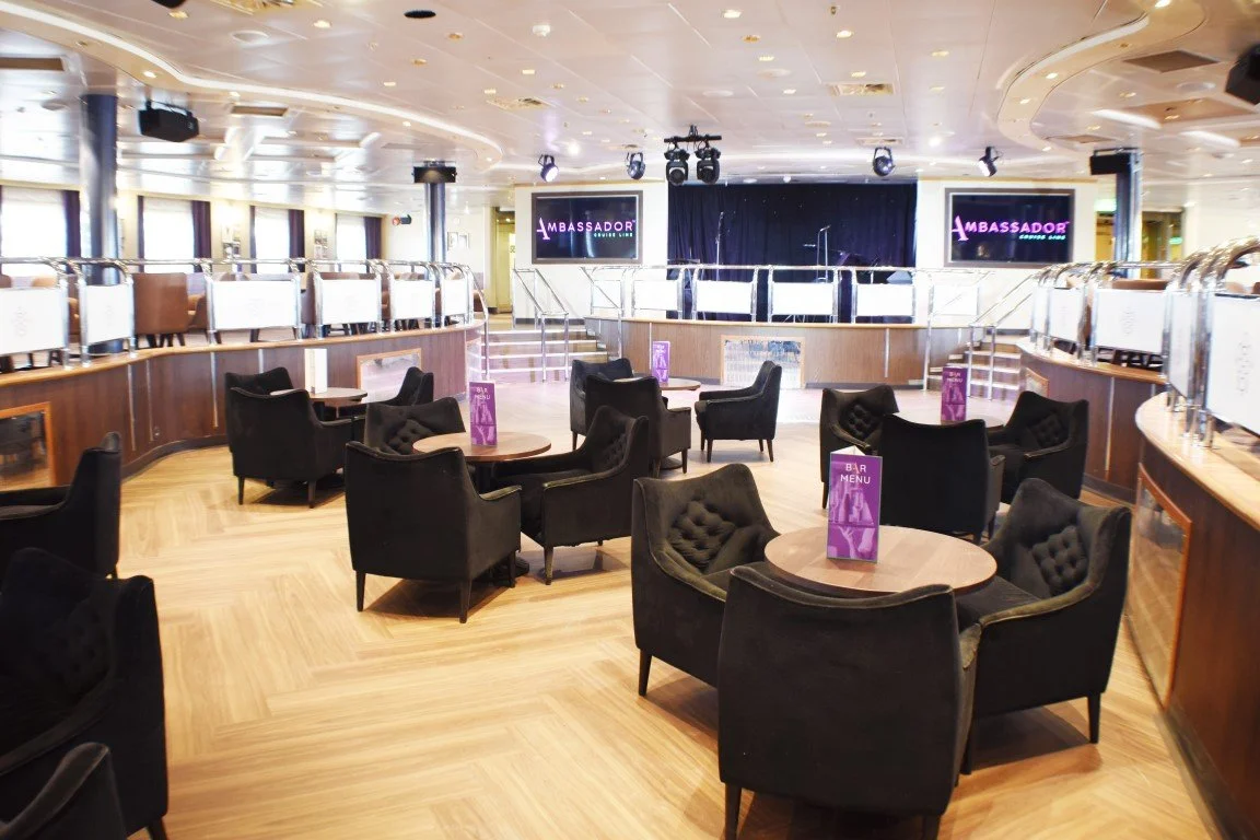 Empty lounge area with black armchairs and round tables, displaying purple bar menus, stage with screens showing "Ambassador", and a dance floor with stage lights in the background.