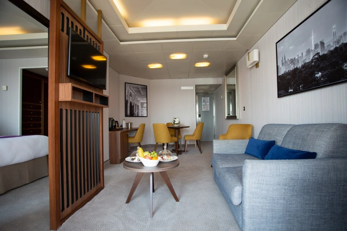 Interior of a modern hotel room with a gray sofa, yellow chairs, and a small coffee table with fruit; wall art, a flat-screen TV, and a ceiling with recessed lighting.