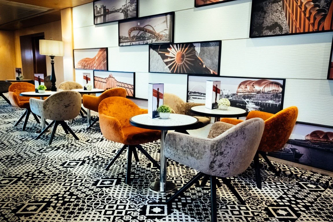 A modern cafe or lounge area with framed cityscape photographs on the wall, several round tables with small plants and menus, and colorful velvet chairs with patterned black and white flooring.