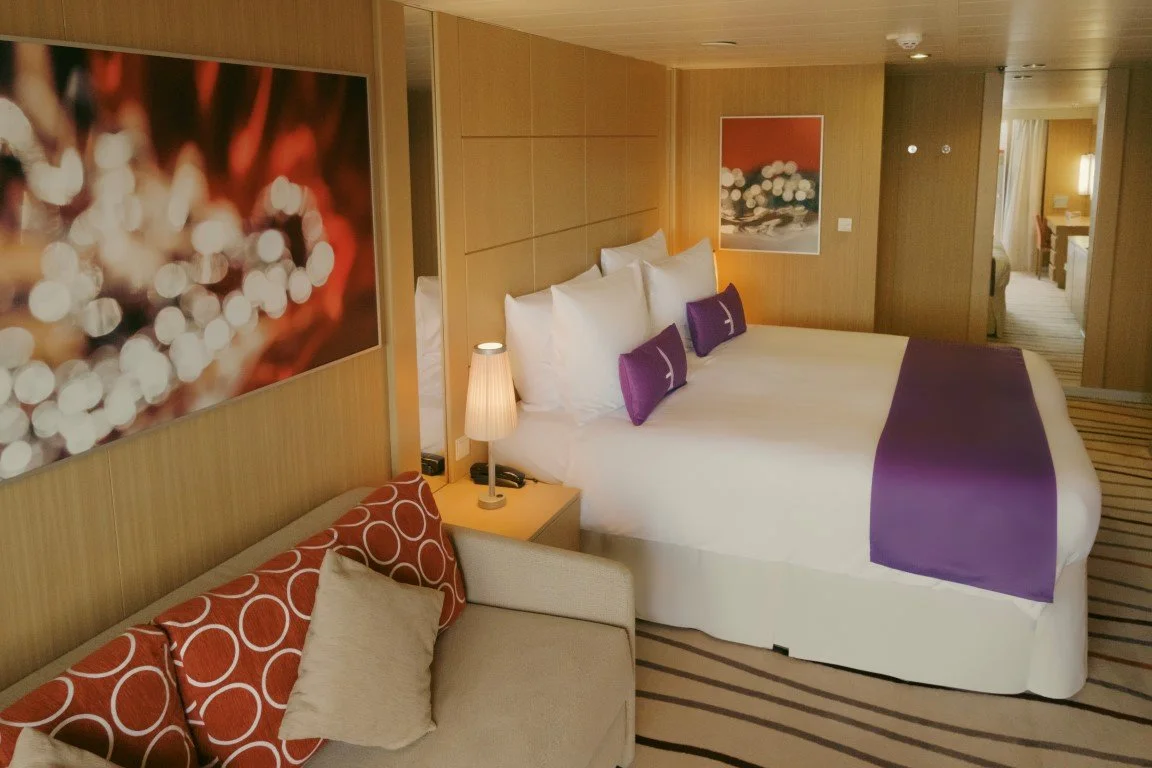 A hotel room with a bed, purple and white bedding, and a sofa with two red pillows with circle patterns. The room has wooden walls, framed artwork, a bedside lamp, and a striped carpet.