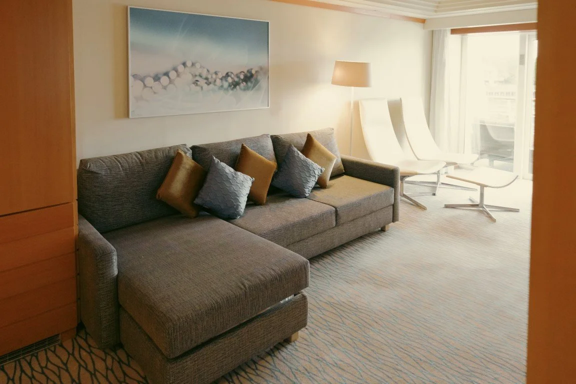 Living room with gray sectional sofa, decorative pillows, a floor lamp, two white modern chairs, a sliding glass door with curtains, a painting on the wall, and patterned carpet.