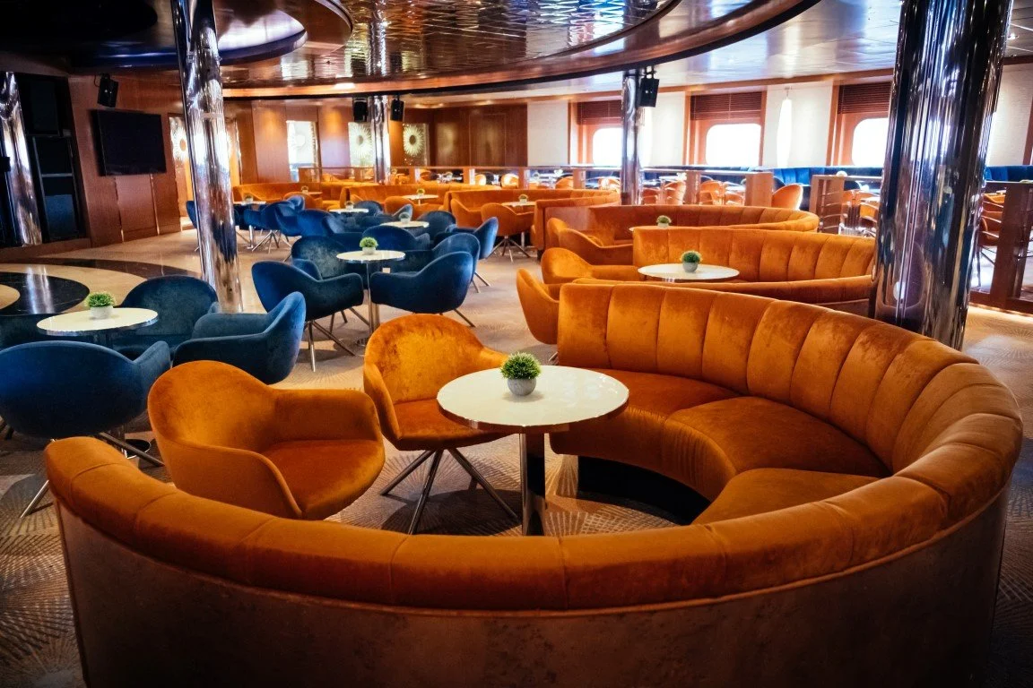 Lounge area with curved orange velvet sofas, small round white tables with small green potted plants, and blue armchairs, on a wooden deck with large windows and wooden paneling.