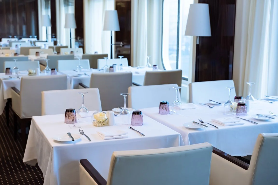 A dining area with white tablecloths, white chairs, wine glasses, and table settings in a well-lit room with large windows and curtains.