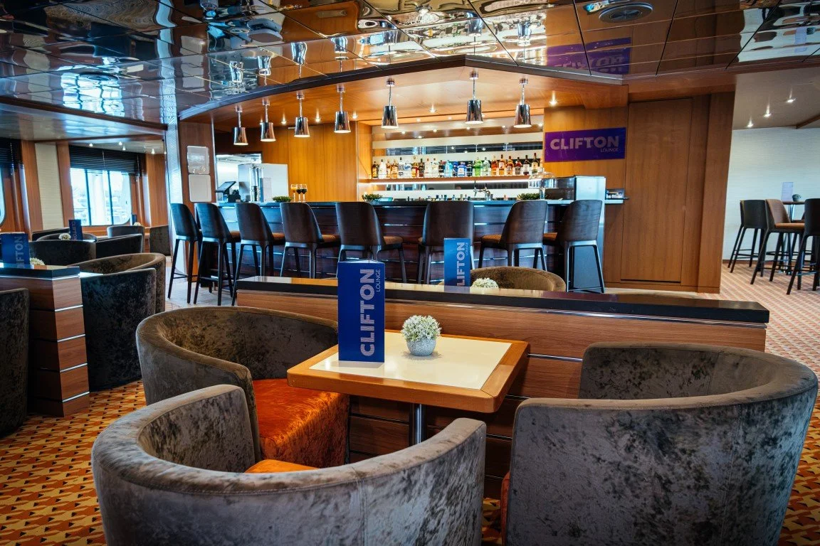 Interior of the Clifton Lounge with a bar area and seating. Bar counter with barstools, shelves with bottles, and a purple Clifton Lounge sign. Plush chairs and tables with blue signage and small plants in a modern, well-lit space.