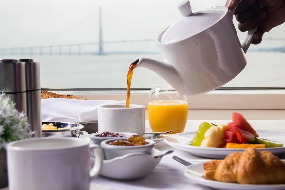 Pouring orange juice into a cup during breakfast with croissants, fruit, and jams, with a window view in the background.