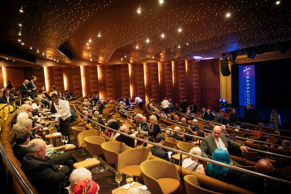 Audience seated and mingling in a theater or conference hall with a phase on the right displaying the word "AMBASSADOR." The ceiling has a star-like lighting pattern, and the room has warm lighting with wood-paneled walls.