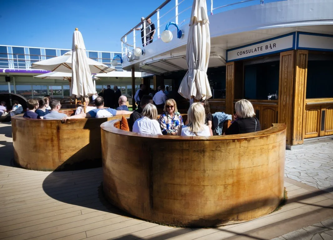People sitting outside at a bar on a cruise ship, with umbrellas and a sign that reads 'Consulate Bar'.