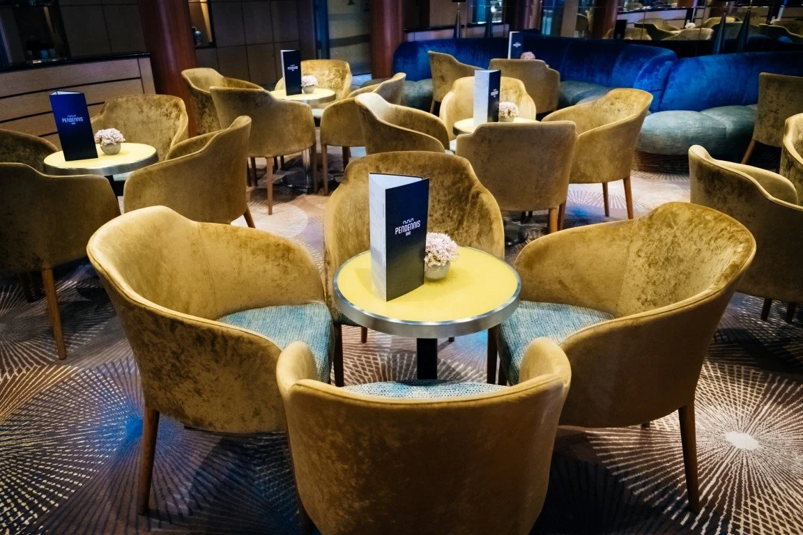 A lounge area with several yellow velvet armchairs and matching small round tables, each with a navy menu and a small white flower arrangement.