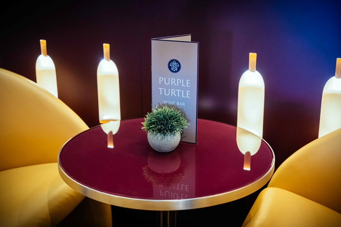 A small round table with a red top and gold trim, flanked by two yellow chairs, with a menu, potted plant, and illuminated bottles in the background, at the Purple Turtle Wine Bar.
