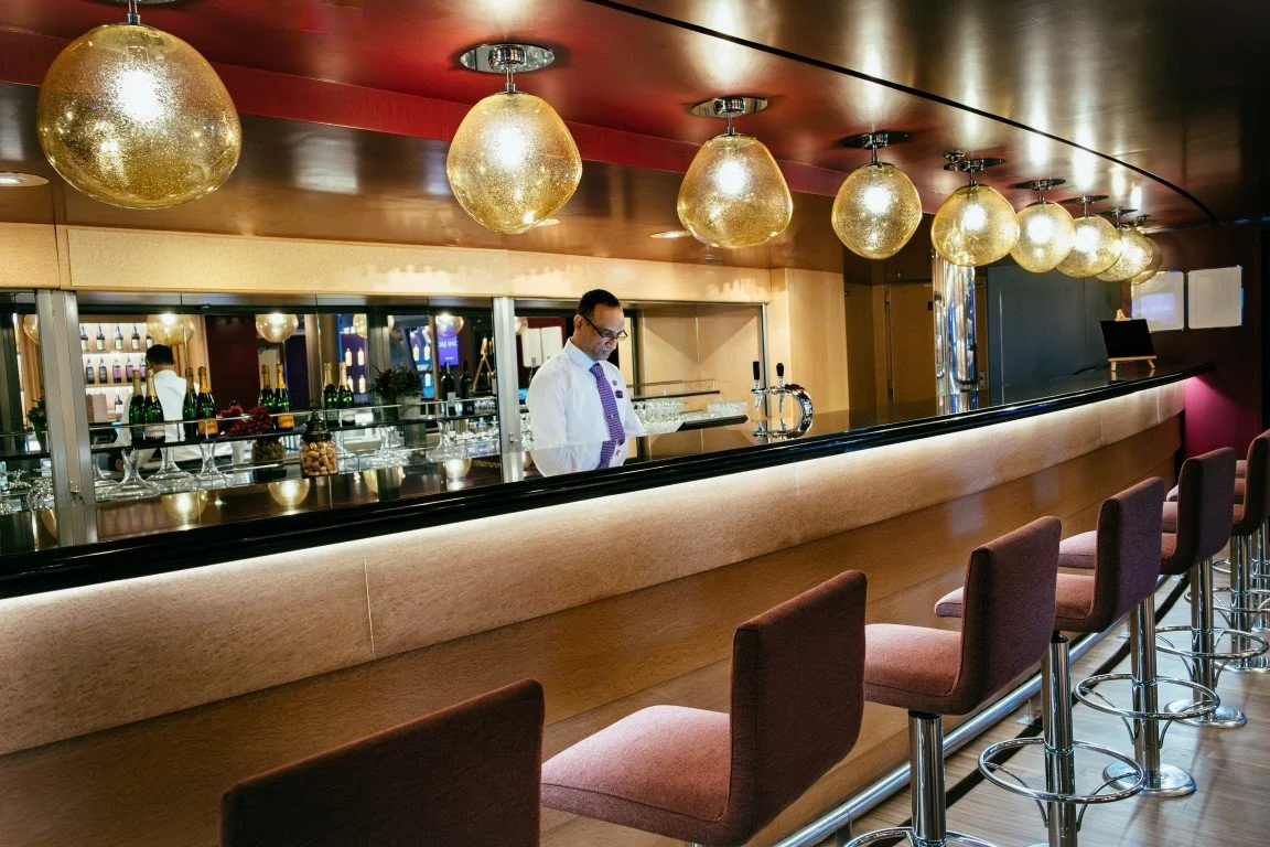 Interior of a modern bar with a person bartender preparing drinks behind the counter, illuminated by large gold spherical pendant lights, with bar stools lined up in front.