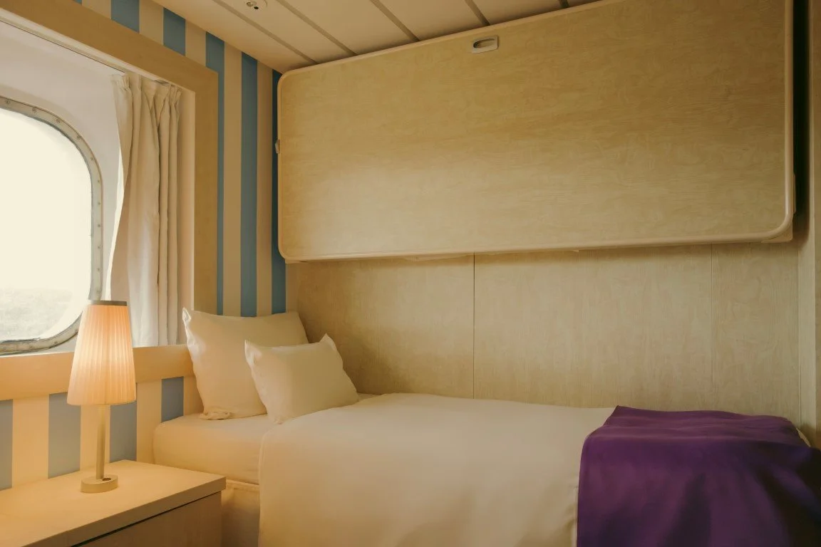 A small bedroom with a bed, purple blanket, bedside lamp, window with curtains, and a folded foldable table or bed platform mounted on the wall above the bed.