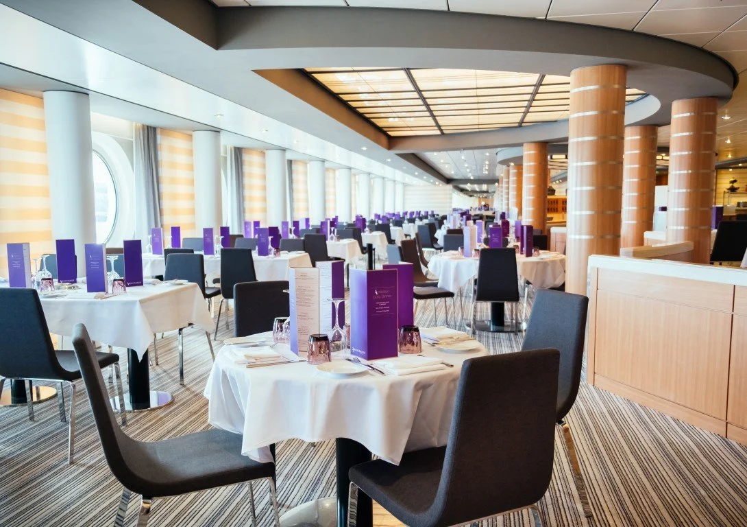A spacious and elegant restaurant with round tables covered with white tablecloths, set with glassware and menus. The restaurant features large windows with curtains, circular columns, and a modern ceiling design with lighting.