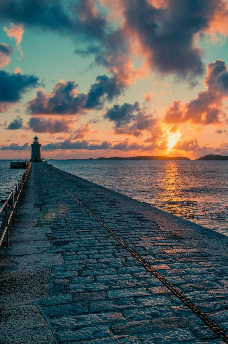 A sunset over the ocean with a lighthouse at the end of a stone pier extending into the water, and colorful clouds in the sky.
