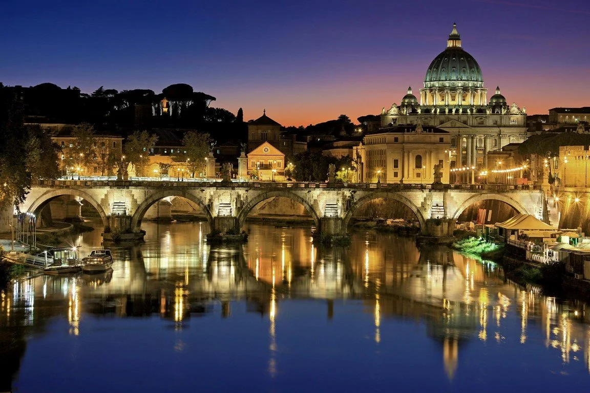 Nighttime view of Vatican City with St. Peter's Basilica illuminated, reflected in the Tiber River, with the city skyline and a bridge in the foreground.