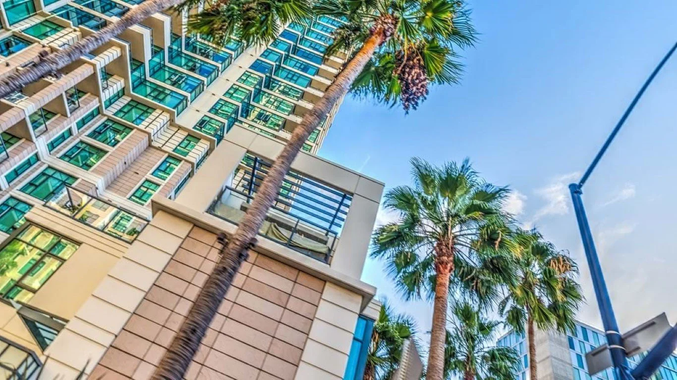 Looking up at tall modern buildings and palm trees against a bright blue sky in an urban area.