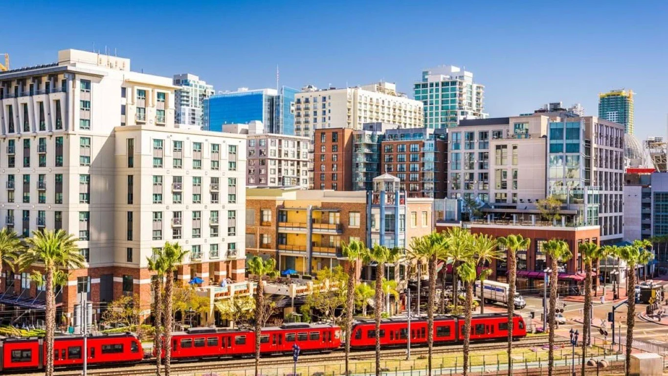 Cityscape with modern buildings, palm trees, and a red train on tracks in the foreground.