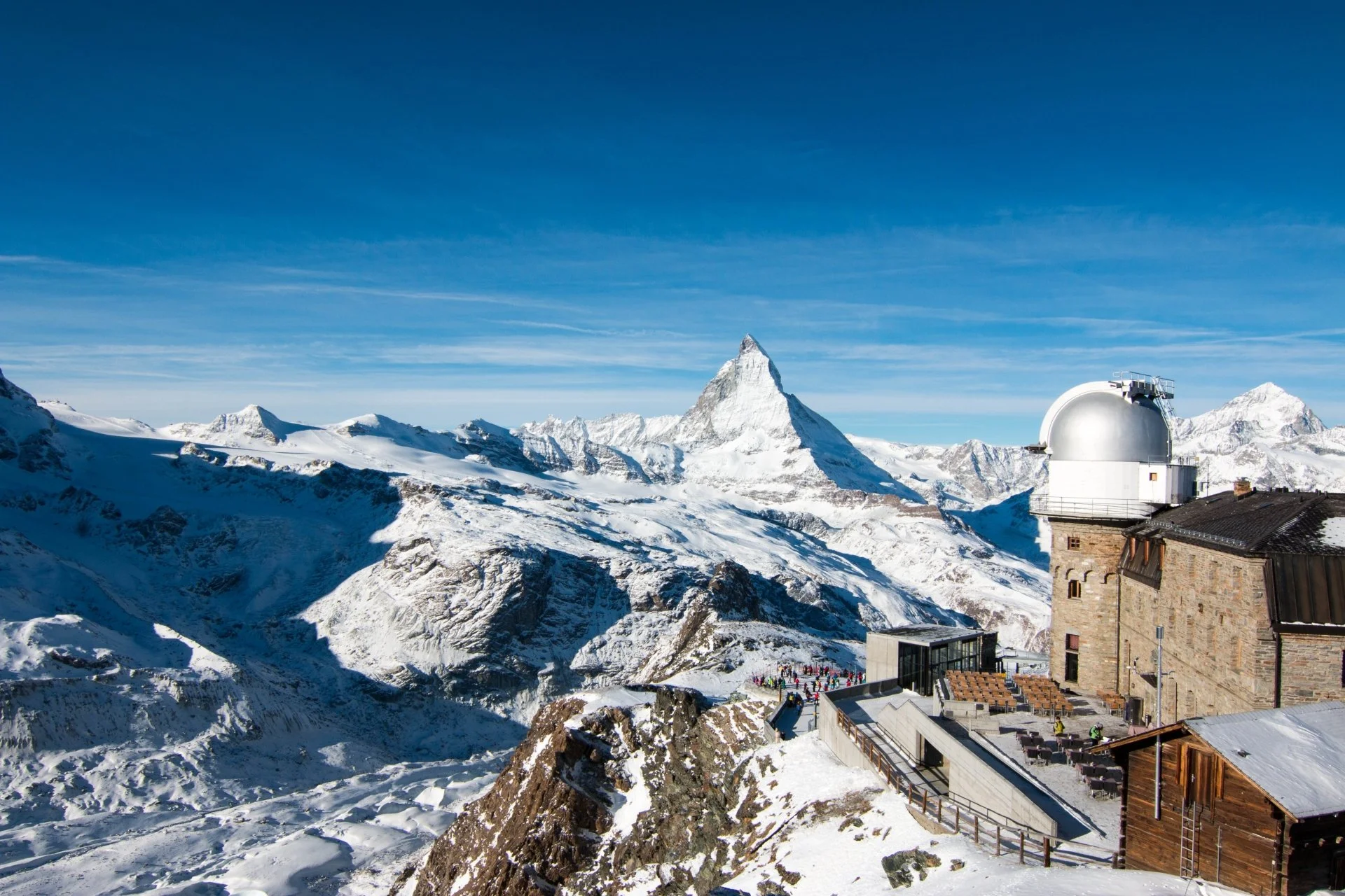 Snow-capped mountains with a prominent peak and an observatory with a white dome on a building, under a clear blue sky.