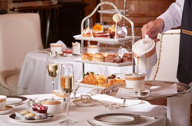 A server pouring tea or coffee into a cup at an elegant high tea setting with tiered trays of pastries, sandwiches, and desserts on a white tablecloth.