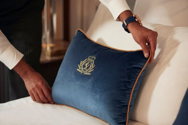 A person placing a decorative blue pillow with gold embroidery on a bed.