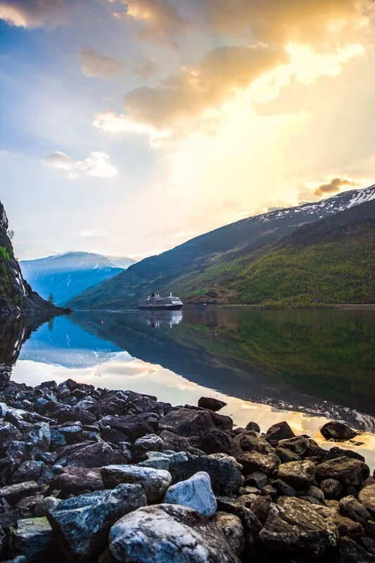 A serene fjord landscape with calm, reflective water, rocky shoreline in the foreground, mountains covered in greenery, and a cruise ship in the distance, under a partly cloudy sky with soft sunlight.