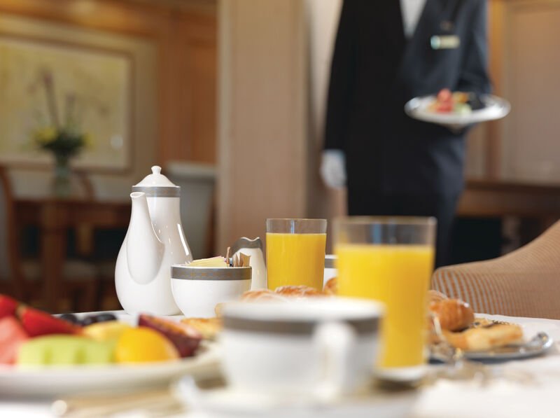 Breakfast table with two glasses of orange juice, a white teapot, a cup, and a plate of assorted fruit and pastries, with a person in formal attire holding a plate in the background.