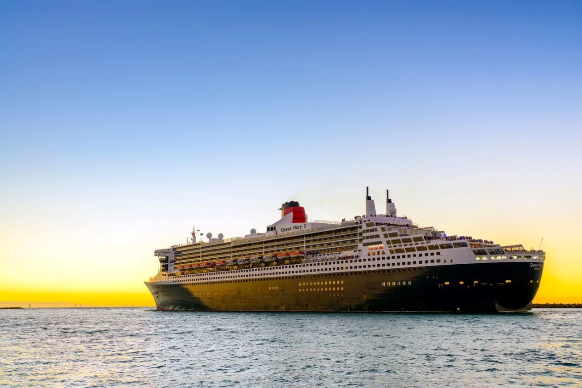 Large cruise ship named Queen Mary 2 sailing on calm water at sunset with a clear sky.