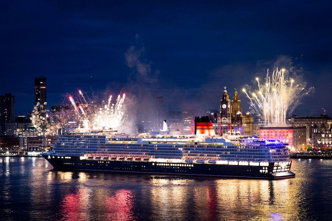 A large cruise ship at night on the water with fireworks lighting up the sky behind. The city skyline with illuminated buildings is visible in the background.