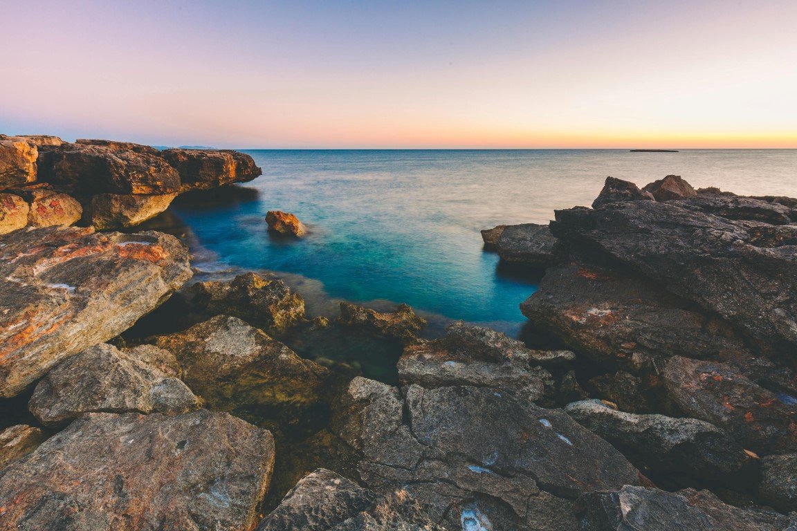 Rocky coastline with calm ocean water and a colorful sunset sky.
