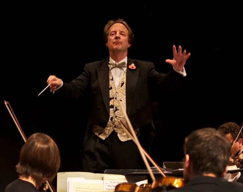 Conductor leading an orchestra during a performance, with musicians playing string instruments.