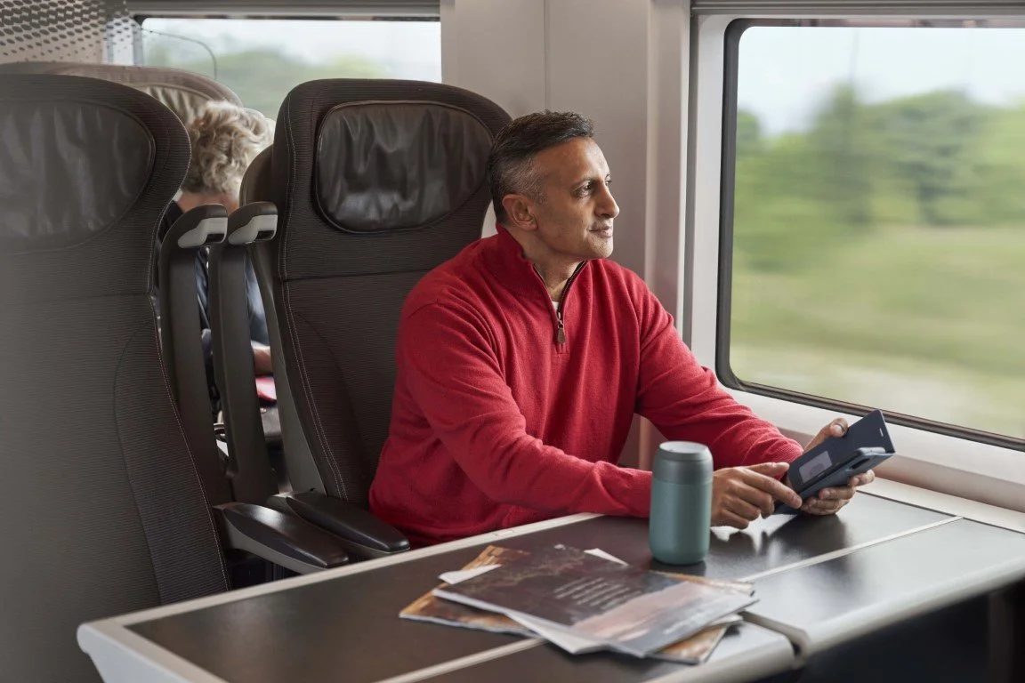 A middle-aged man with short dark hair and a red zip-up sweater sitting in a train seat, looking out the window and holding a smartphone. A woman with gray hair is seated behind him, partially obscured. The table in front of him has magazines and a blue travel mug.