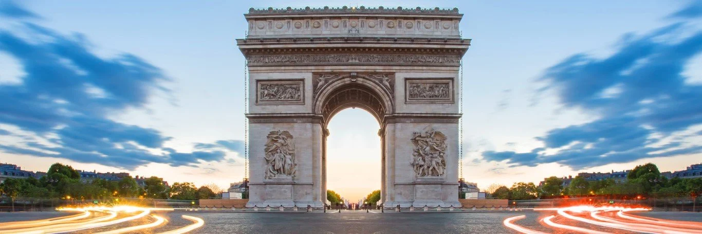 The Arc de Triomphe in Paris, France, with light trails from moving vehicles on the street and a sky with scattered clouds during sunset or sunrise.