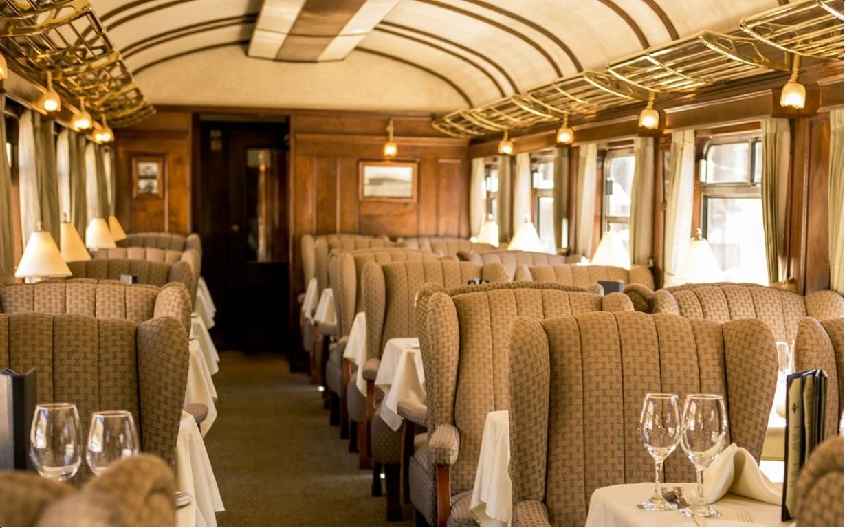 Interior of a vintage train dining car with wooden walls, patterned upholstered seats, tables set with white tablecloths, wine glasses, and covered with small lamps, with windows along sides.