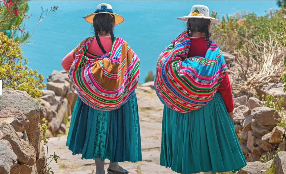 Two women dressed in traditional Bolivian clothing, wearing colorful shawls and wide-brimmed hats, standing on a trail overlooking a lake.