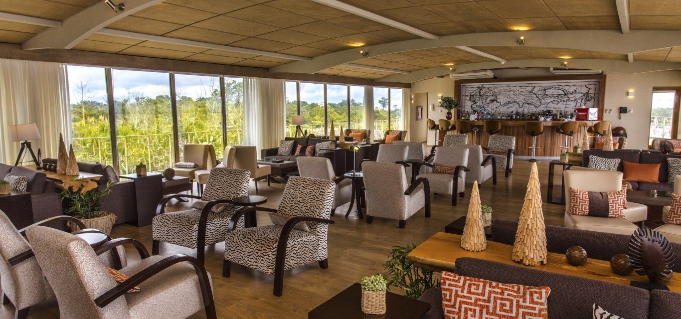 Interior of a spacious lounge with large windows, various armchairs and sofas, a bar area in the back, and decorative objects on tables.