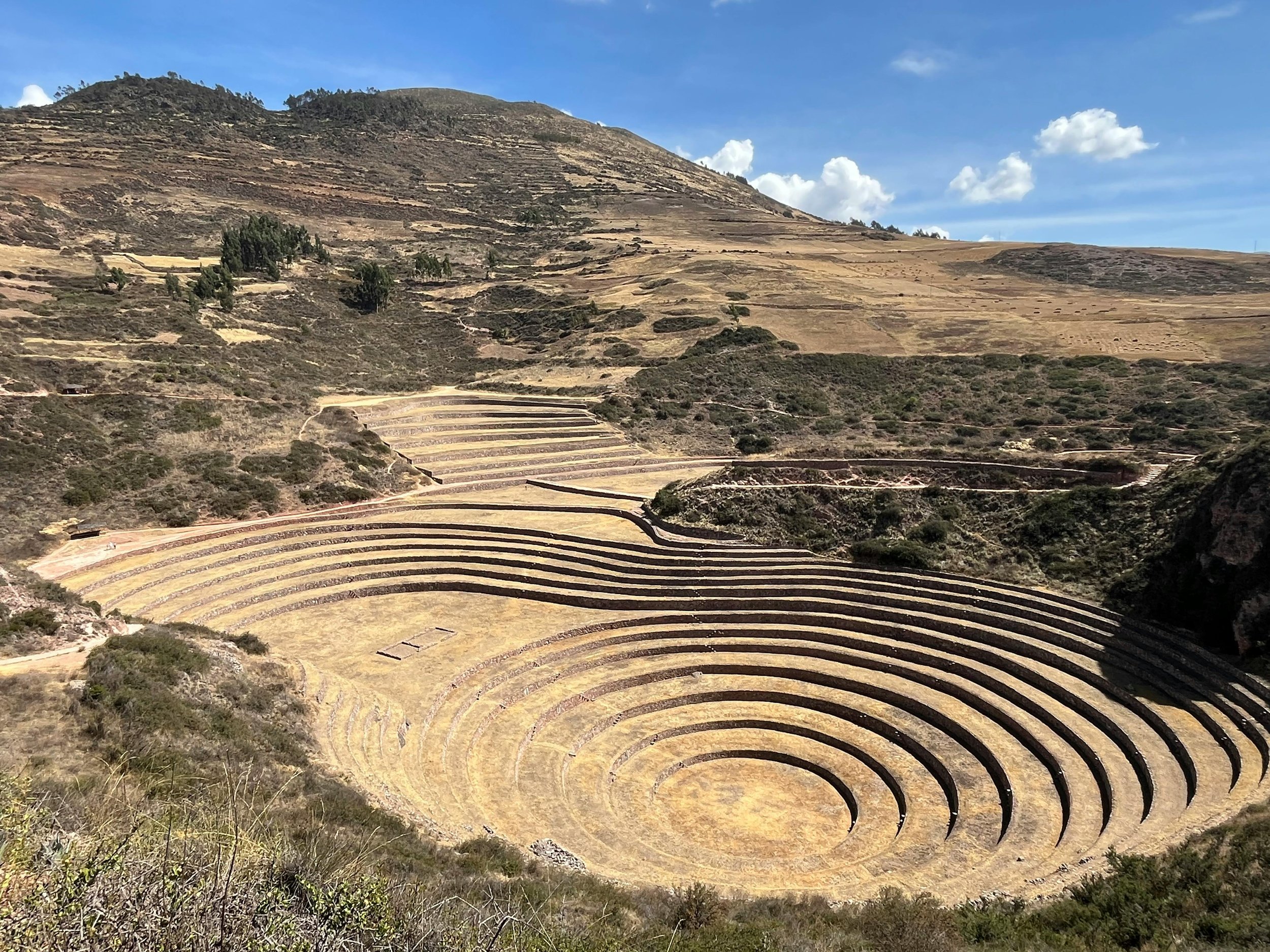 Ancient Incan archaeological site with terraced circular structures in a mountainous landscape under a blue sky with clouds.
