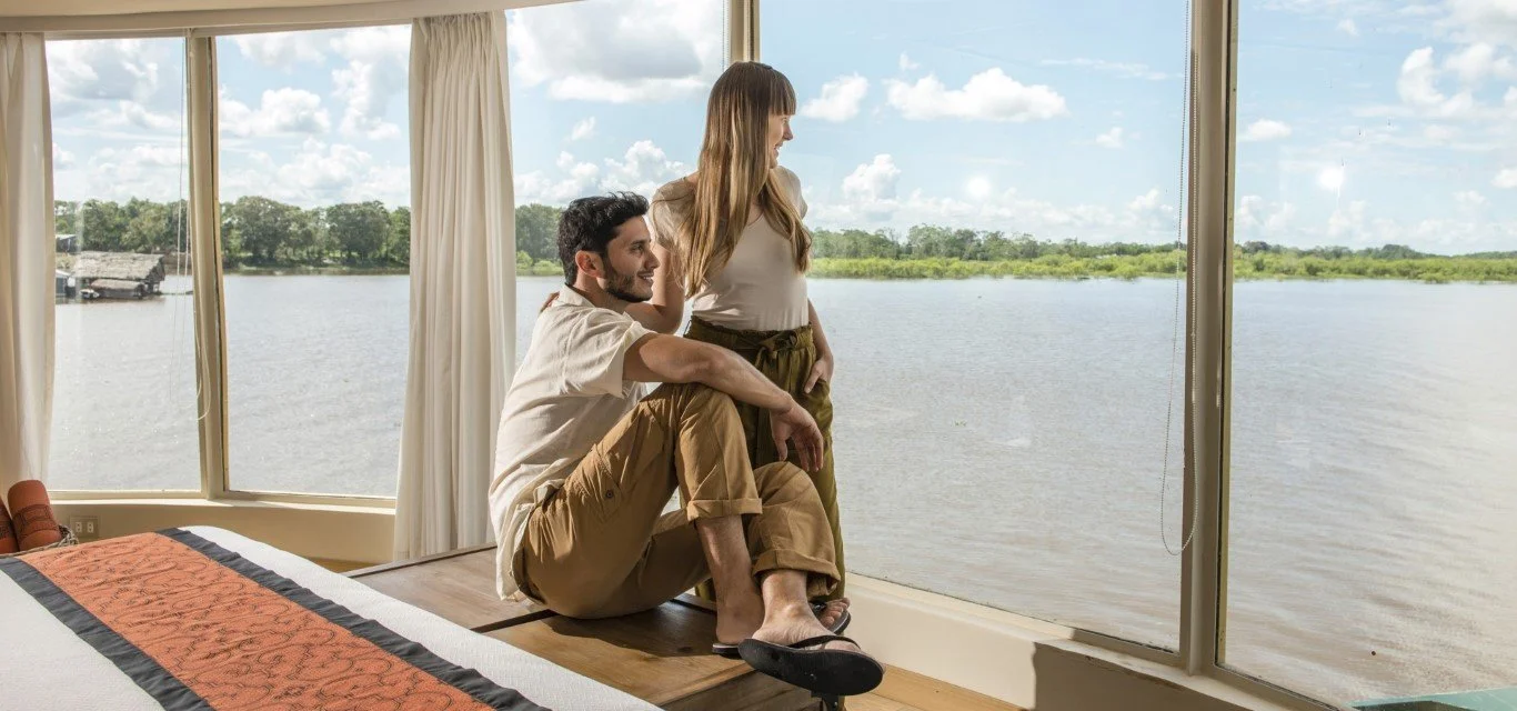 A man and a woman inside a room with large windows overlooking a river and a forest, with the man sitting on the floor and the woman standing.
