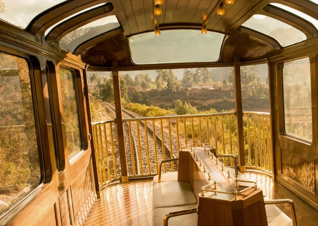 View from the front of a vintage train with wooden interior and golden railings, traveling through a lush, green landscape.