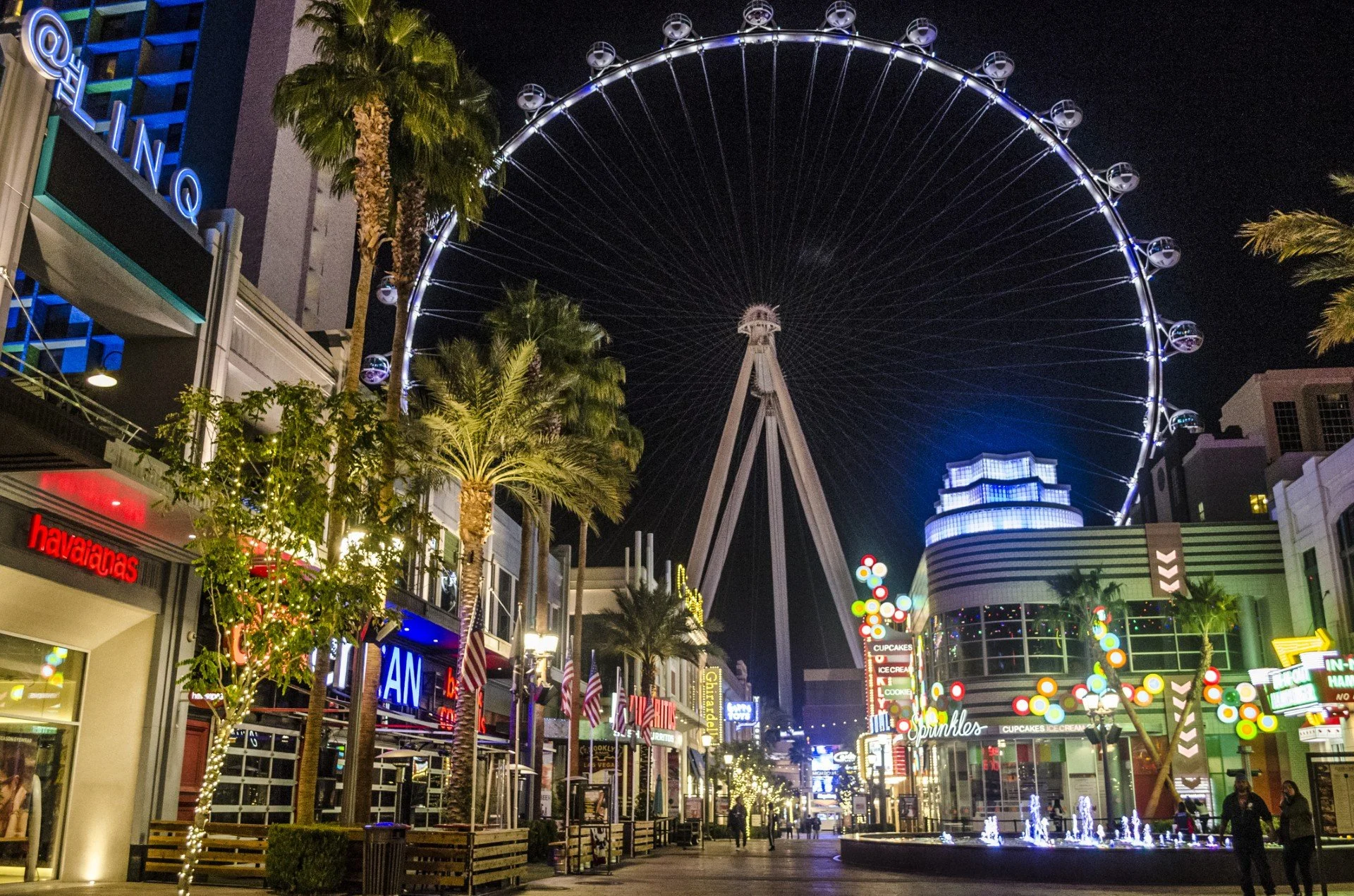 Night view of a busy outdoor shopping and entertainment area with a large illuminated Ferris wheel in the background, palm trees decorated with string lights, storefronts with bright signs, festive decorations, and people walking along the street.