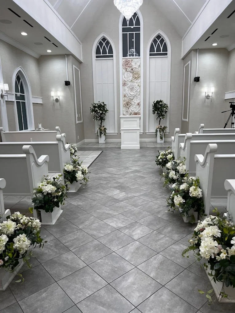 Empty wedding chapel with white pews decorated with white flowers, leading to an altar with floral decorations and tall windows.