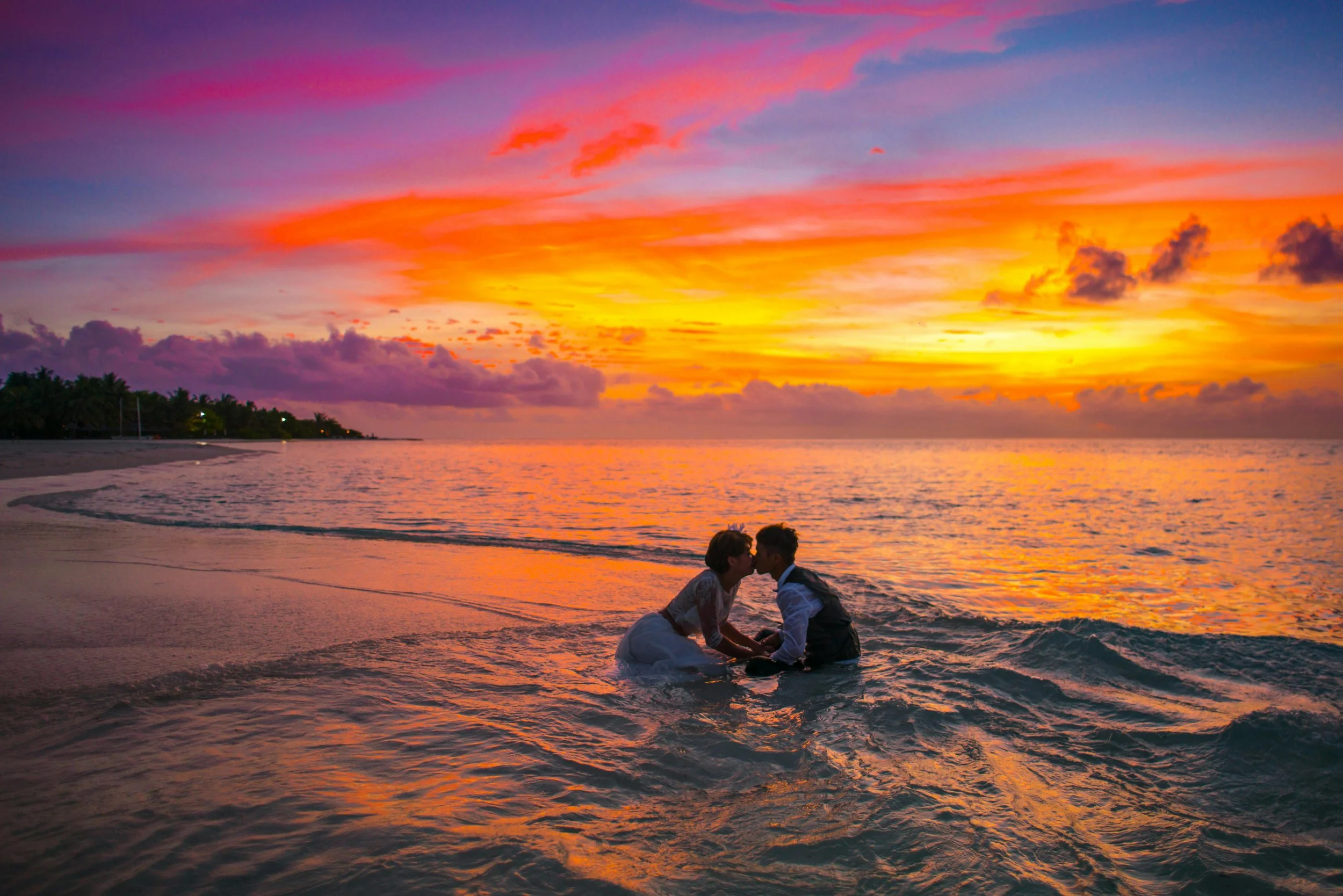 A couple in wedding attire sharing a kiss in the ocean at sunset with colorful sky and horizon in the background.