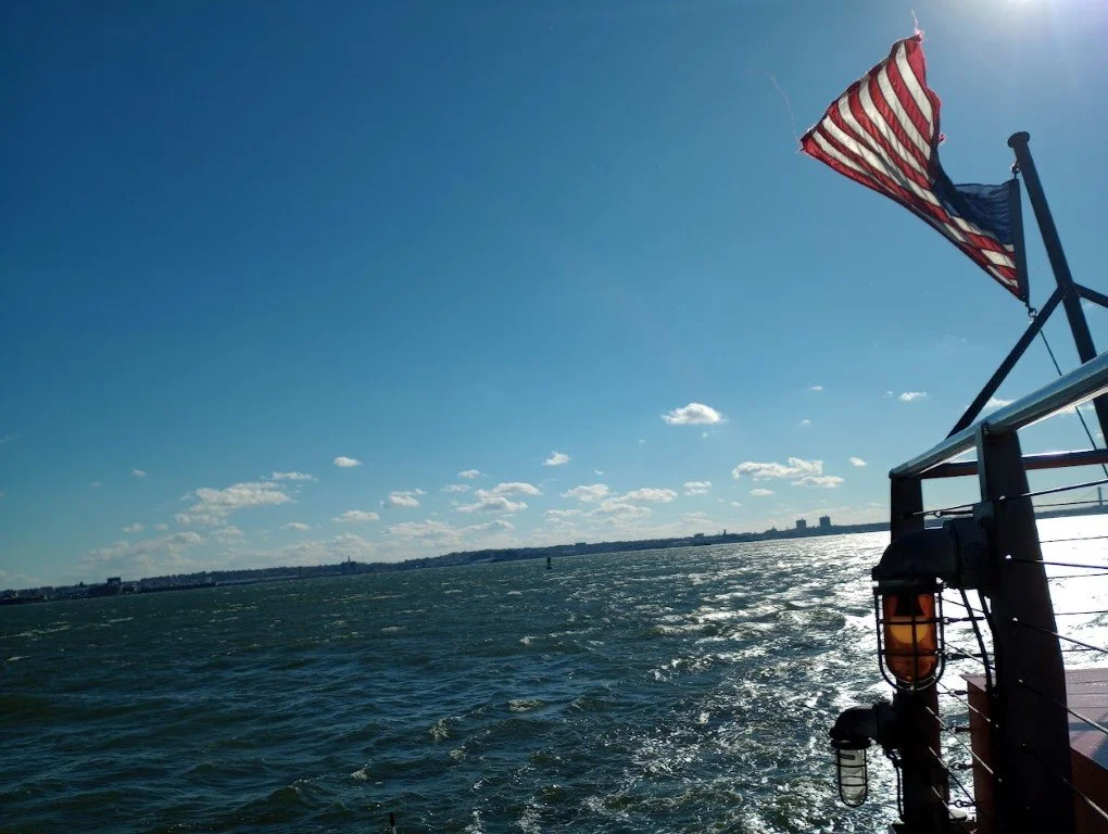 View from a boat with an American flag waving in the wind, water splashing below, and a distant skyline on the horizon under a sunny sky with scattered clouds.